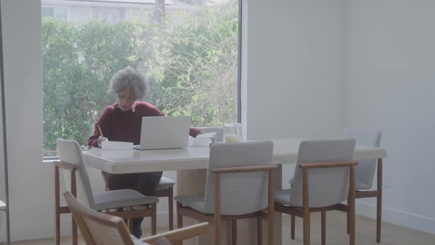 A woman with curly gray hair works at a bright dining table with a laptop and notebook surrounded by minimalist decor and natural light