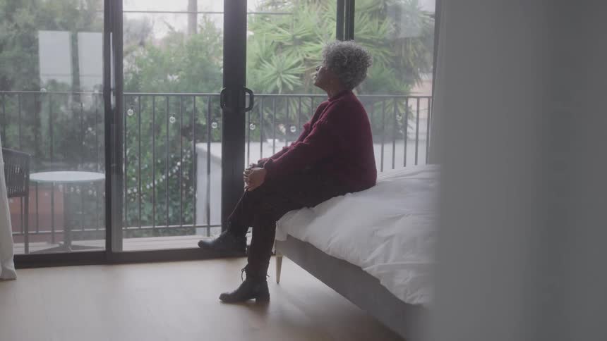 An elderly woman sits alone on a bed in the bedroom at midday wearing black jeans boots and a red sweatshirt with her legs crossed and trees visible outside through the glass window
