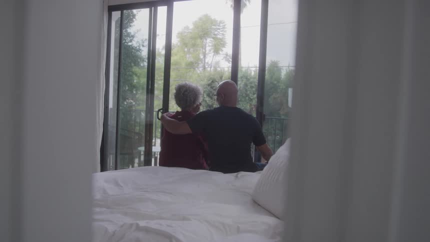 An older couple sits closely on the edge of a bed quietly gazing out a large window framed by white curtains