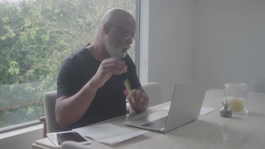 A man sits at a bright kitchen table with a laptop holding papers and a pencil possibly reviewing documents or plans