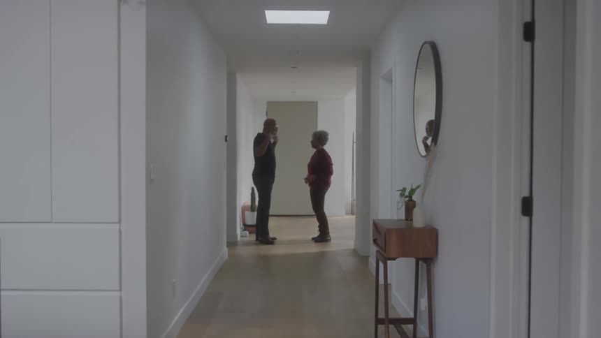 An adult man and an older adult woman stand facing each other in a bright modern hallway with light colored walls and wooden floors conversing with hand gestures as a mirror and a small table with a plant are visible to the right