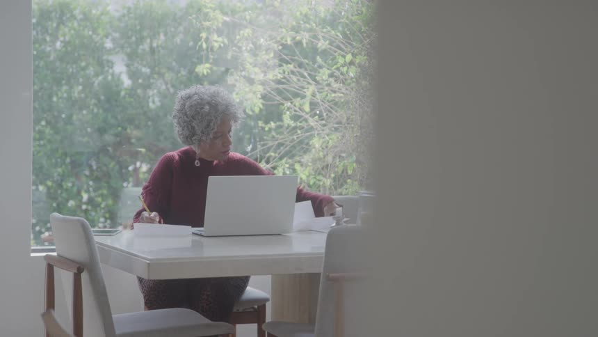 Elderly woman writing with a pen while sitting at a desk with an open laptop