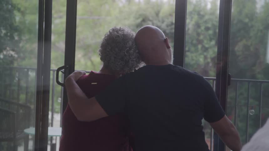 An elderly black man hugs his female partner as they both sit in a room during daytime