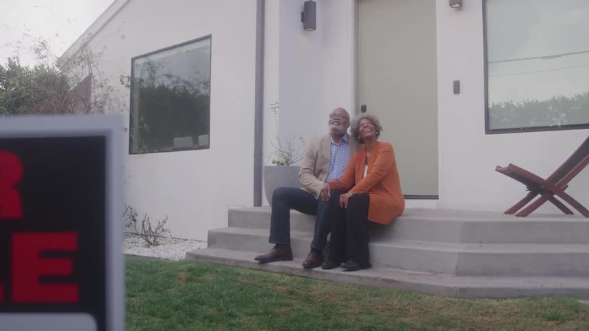 An elderly couple sits on the front porch of a house hugging each other with excitement while a for sale sign stands on the lawn under the midday sun