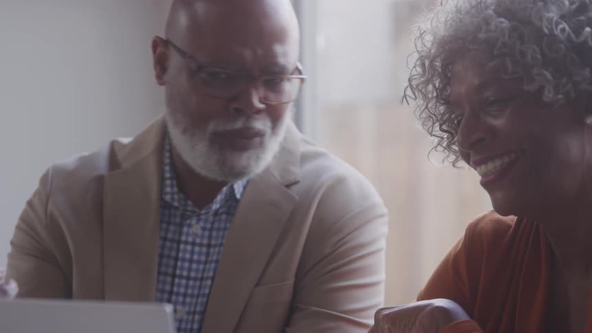 A man smiles while watching a woman point at something on a laptop both appearing engaged and amused