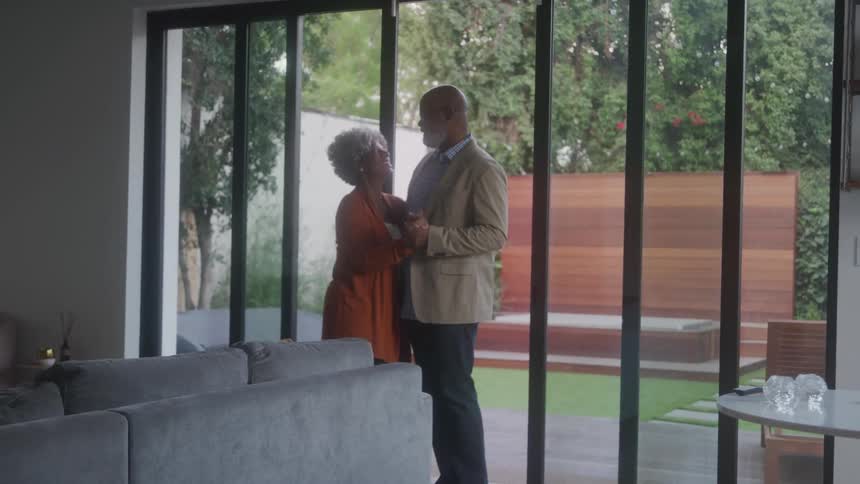 An affectionate older couple shares a dance by the glass doors of a cozy modern living room overlooking a lush backyard