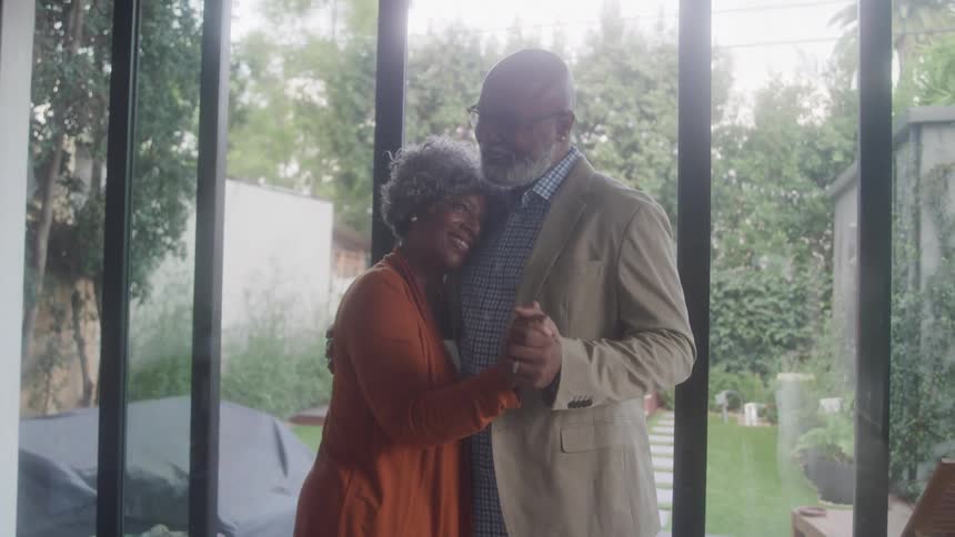An elderly couple the man in a light colored jacket and the woman in an orange dress dance slowly holding hands and embracing each other in front of large glass doors that open to a lush green backyard