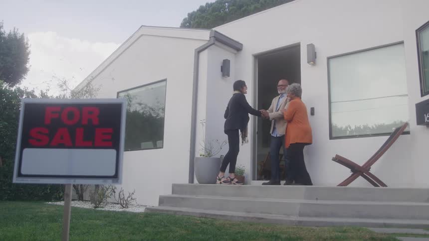 An older adult couple stands smiling on the porch of a modern white house as a young adult female real estate agent hands them keys to their new home with a red for sale sign on the lawn