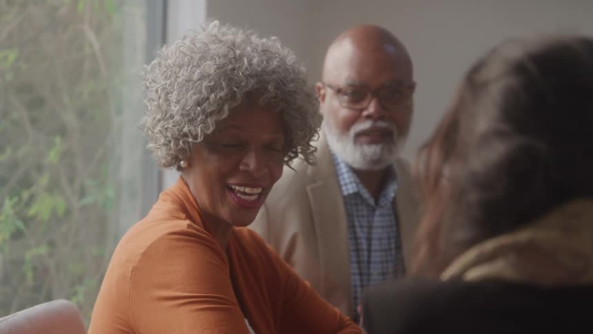 An older couple sits attentively at a meeting table engaged in conversation with someone seated across from them in a bright room