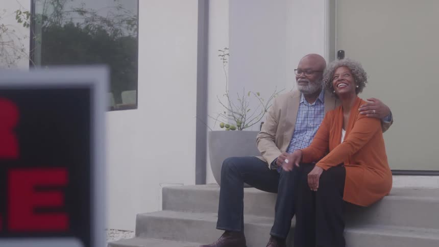 Older couple sits on the front steps cuddling and smiling while a for sale sign stands in the front yard