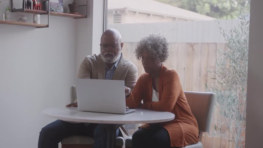 A couple sits closely at a small round table collaborating while looking at a laptop screen in a cozy sunlit room