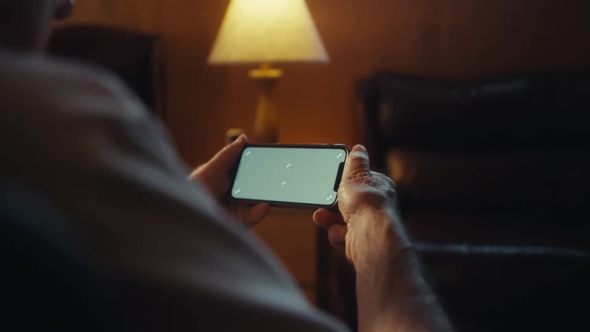 An older sad looking man holds a smartphone horizontally engrossed in its screen while seated in a cozy warmly lit living room