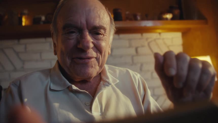Elderly man sitting in a chair at home gestures during a video call on his smartphone
