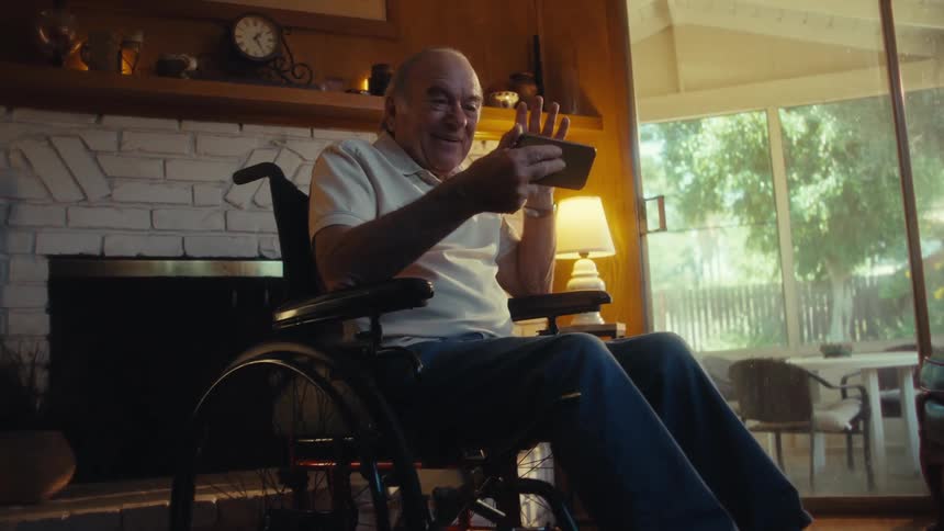 A joyful elderly man in a wheelchair with a white polo shirt and jeans is enthusiastically engaging with a smartphone in a sunlit living room with a brick fireplace and large windows