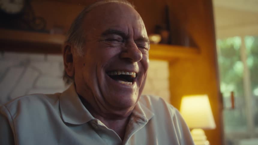 This video features a closeup of an older man with light brown hair dressed in a lightcolored polo shirt laughing heartily as he looks at and interacts with a smartphone the warm indoor lighting and the natural background suggest a comfortable