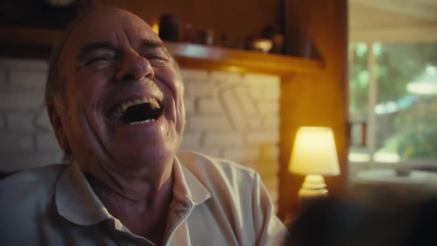 An elderly man suddenly starts laughing while watching a program on his computer while sitting indoors