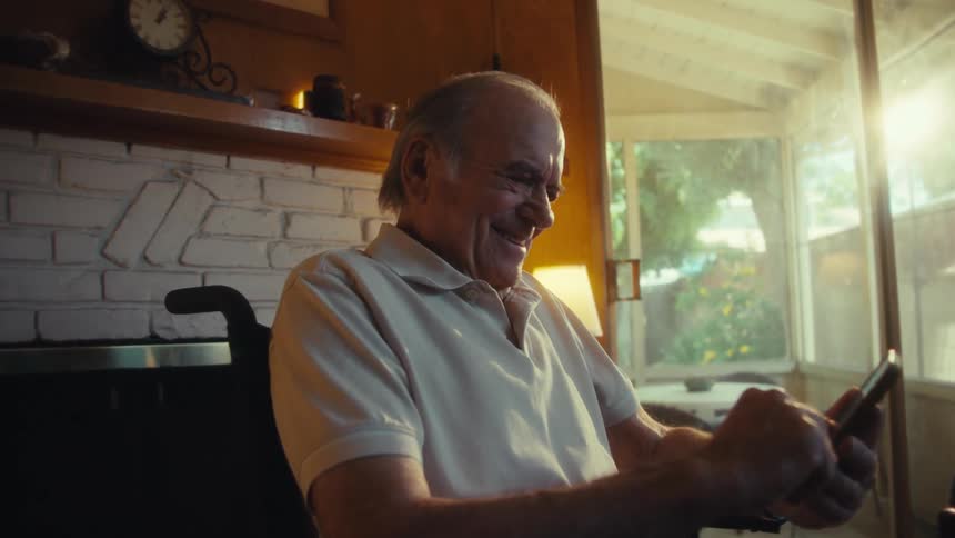 This video shows an older man in a wheelchair comfortably seated in a sunlit living room happily engaging with a smartphone the warm light streaming through large windows suggests a peaceful home environment as he smiles and interacts with the device