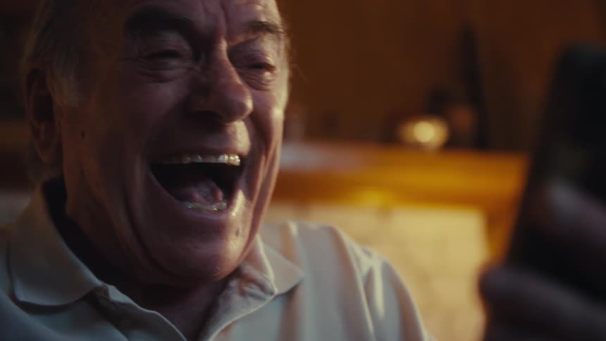 An elderly man sit in a sunlit room using his phone while laughing