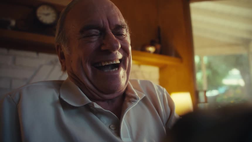 An elderly man laughs wholeheartedly whiling holding a cell phone in warm lighting