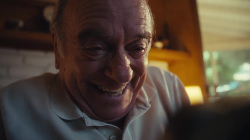 An elderly man with a warm smile and wrinkles around his eyes wearing a lightcolored collared shirt is captured in a closeup shot laughing to his smartphone with a softly lit domestic background