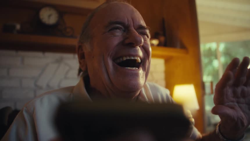 An elderly man smiles warmly in soft natural light while looking at a phone in front of him