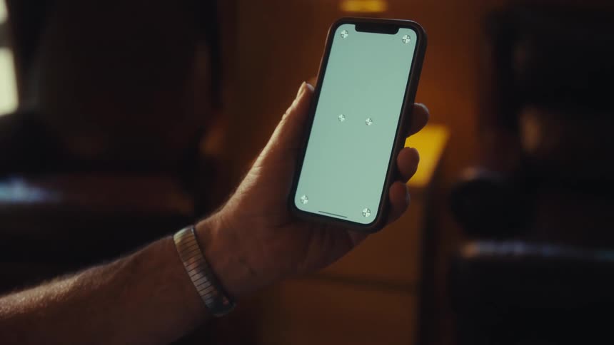 A smiling older man sits comfortably in a warmly lit room holding up a smartphone