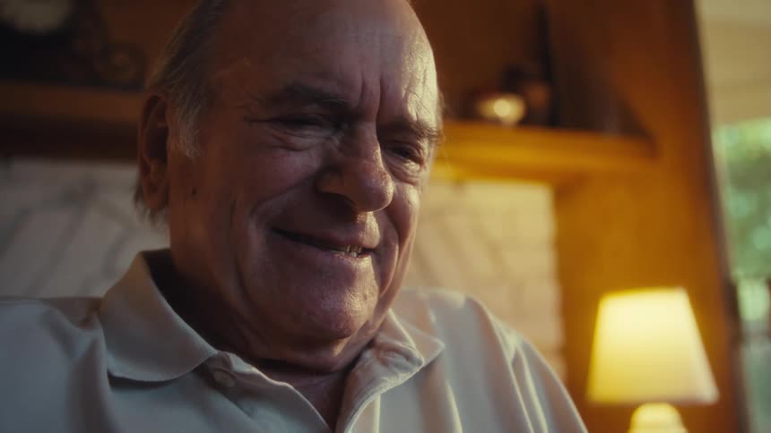 Elderly man sitting in a wheelchair in his living room smiles while scrolling on his smartphone