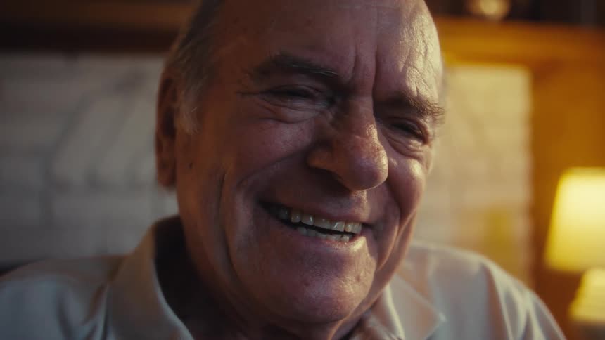 An elderly man wearing a lightcolored polo shirt and jeans is seated in a wheelchair in a sunlit living room with a brick fireplace attentively holding and looking at a smartphone