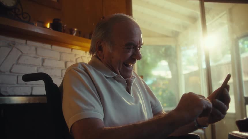 Elderly man sitting in a wheelchair laughing and interacting with a smartphone