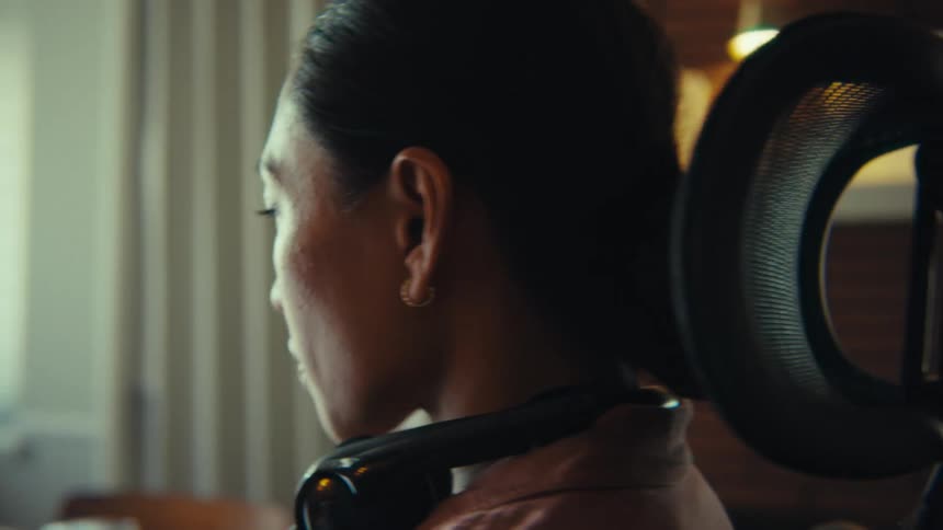 This video features a professional woman likely in her 30s or 40s with dark hair tied back wearing black headphones around her neck and a light pink collared shirt the camera focuses on her face as she looks to the side then shifts to a wider shot