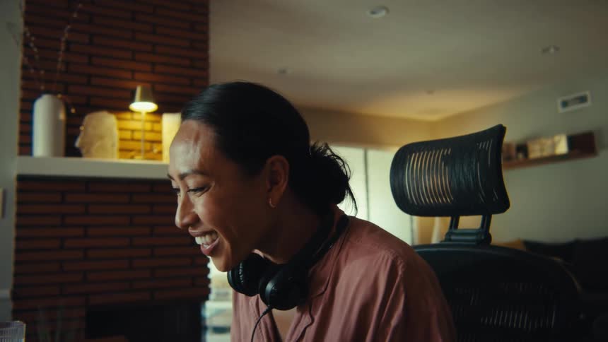 This video features a professionallooking woman with dark hair wearing headphones and a pink top laughing and smiling while working on a laptop the cozy welllit home environment complete with a fireplace and warm lighting