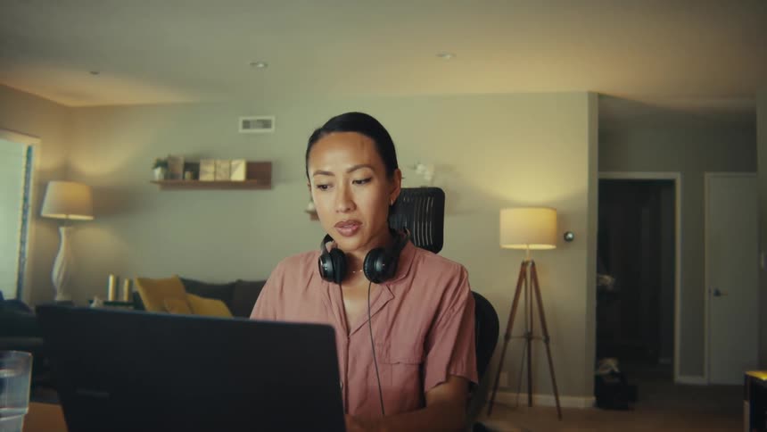 This video features a professionallooking woman with long dark hair and headphones around her neck intently focused on a laptop in a bright modern living room or home office papers and office supplies are neatly arranged on the table in front of her