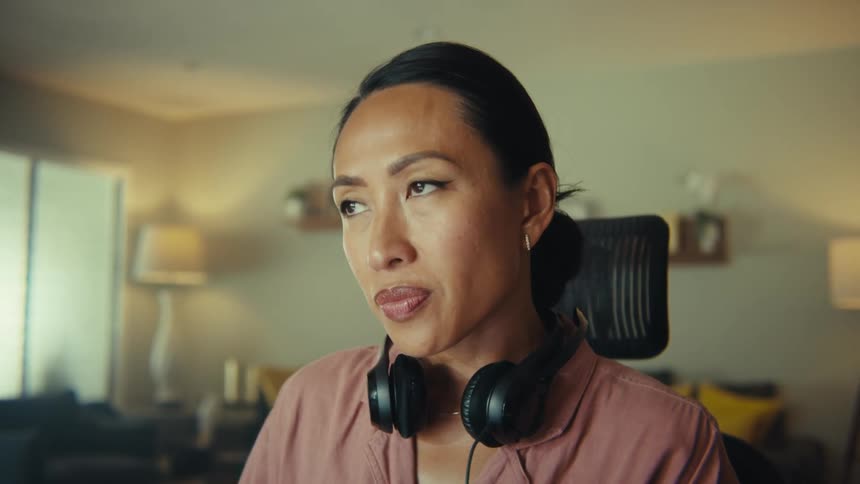 A young woman wearing sits in an office chair in her living room with headphones resting on her shoulders as she operates a laptop she suddenly becomes lost in thought with a couch and two lamps positioned on the left and right sides of the room