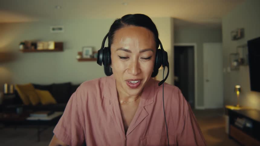 This video features a professional woman likely in her 30s or 40s with dark hair tied back wearing a pink collared shirt and black headphones around her neck she is seated in a modern living room or home office actively engaged in a video call