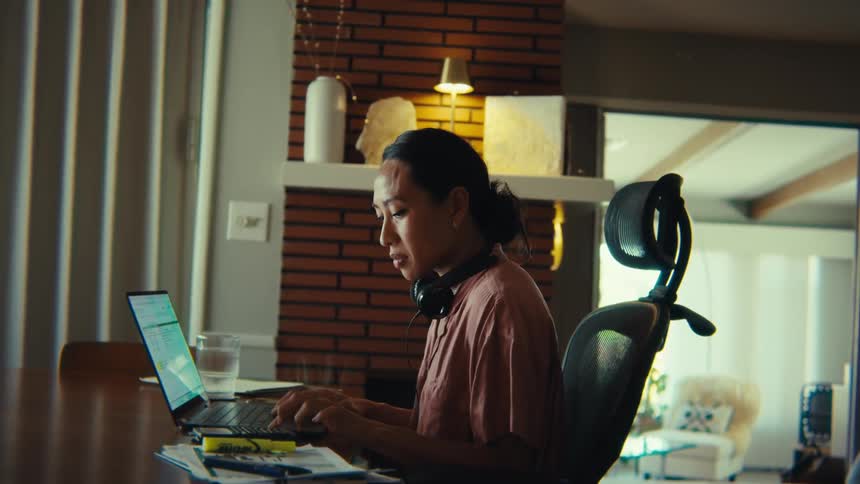 This video shows a focused woman with dark hair wearing headphones diligently working on her laptop at a desk the background features exposed brick a warm lamp and various decorative items creating a cozy yet productive workfromhome atmosphere