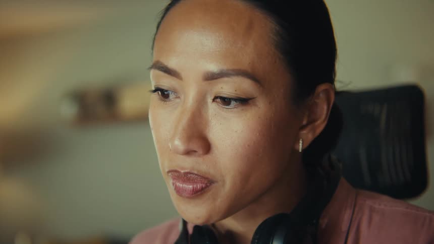A woman with her hair pulled back and headphones around her neck sits at a desk intently focused on her laptop the background reveals a cozy living space with a sofa shelves and a tv suggesting she is working from home