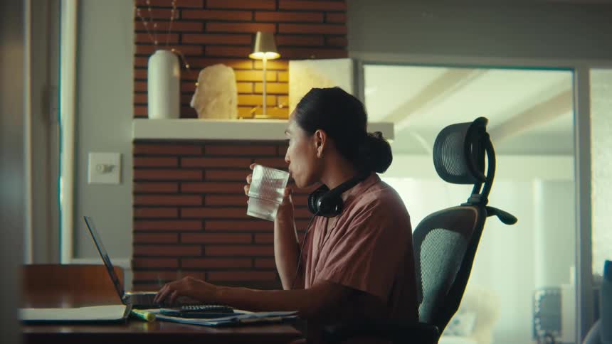 A woman sitting behind a desk in her living room working on a laptop suddenly picks up a glass of water to take a sip with headphones hanging around her neck