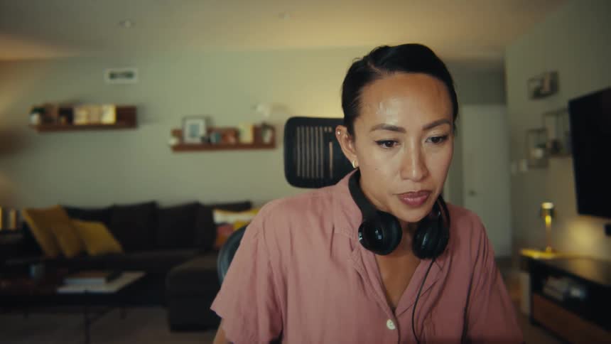 This video features a professional woman likely in her 30s or 40s with dark hair tied back and black headphones around her neck engaged in a focused work session on her laptop the scene alternates between a headshot of her looking at the screen