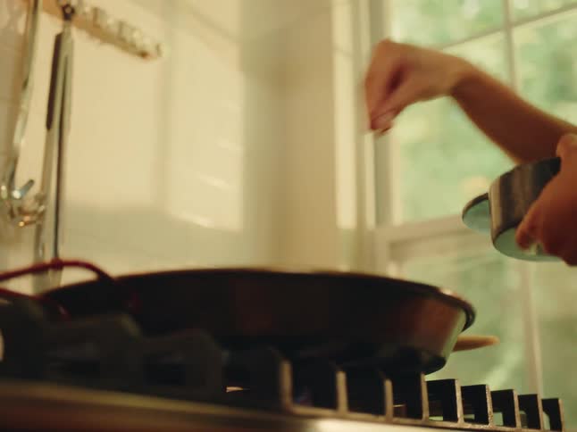 A closeup shot captures a hand sprinkling seasoning into a dark pan on a stovetop with a sunlit window and kitchen items blurred in the background