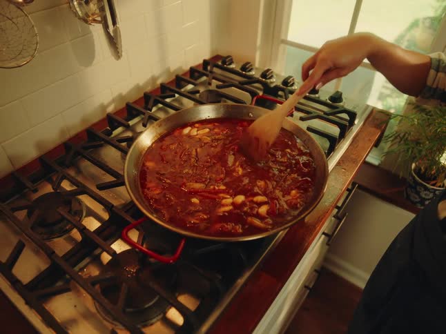 Person with a wooden spoon cooking in a pot on the stove with red sauce