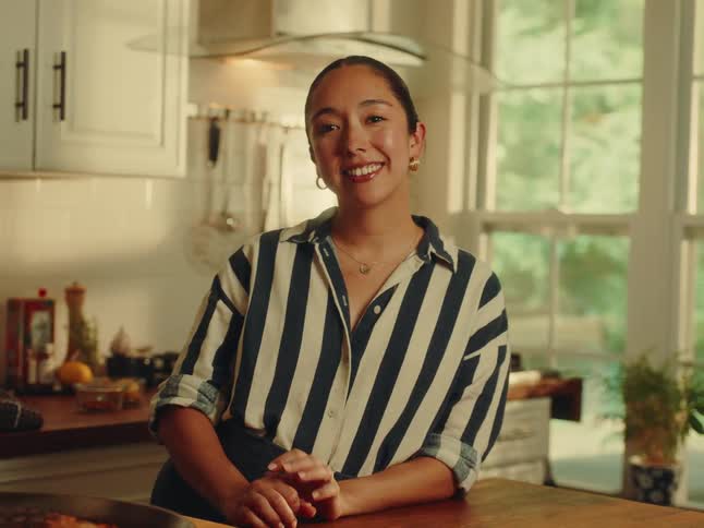 A radiant woman with her hair pulled back wearing a navy and white striped collared shirt beams directly at the camera from a bright welllit kitchen