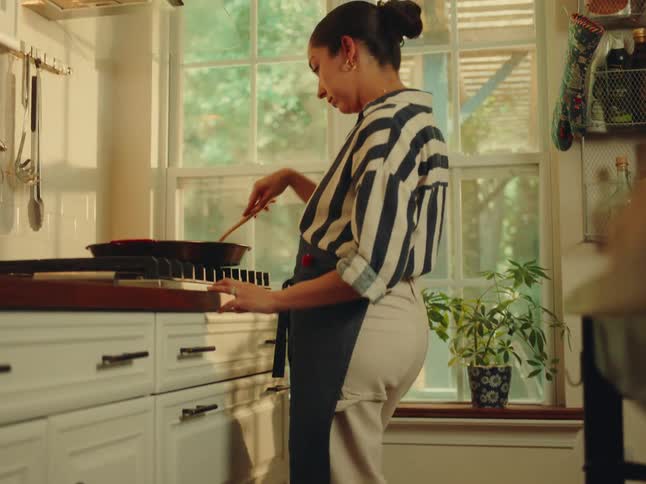 A woman in a striped shirt and dark apron is seen cooking in a kitchen stirring a pan on the stovetop sunlight streams through a large window illuminating the room and a potted plant on the windowsill