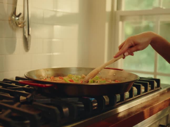 A hispanic woman with her hair in a bun wearing a striped shirt and hoop earrings is cooking at the kitchen and there are kitchen items blurred in the background