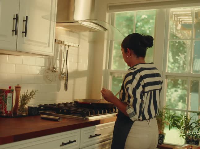 A woman with dark hair tied back wearing a striped shirt and apron is cooking in a bright kitchen she is stirring something in a pan on the stovetop with natural light streaming in from a large window behind her illuminating the rusticstyle kitchen