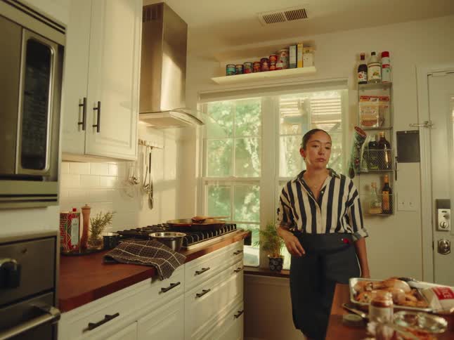 A woman in a striped shirt and apron cooks on a stovetop in a sunlit kitchen then turns to prepare ingredients on a nearby counter