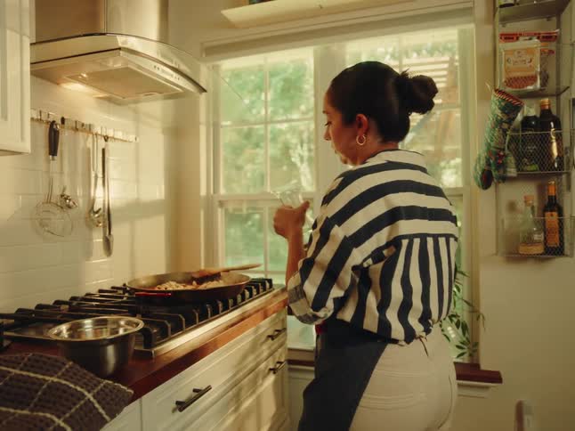 A woman wearing a striped shirt and an apron is seen cooking on a gas stove adding ingredients and pouring liquid from a glass decanter into a large pan