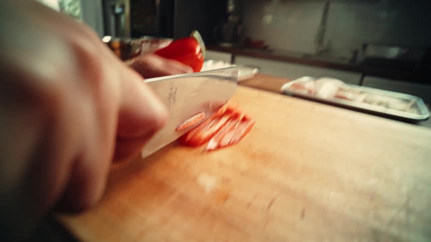 Hand is holding a knife and is slicing into a red bell pepper while using a wood cutting board