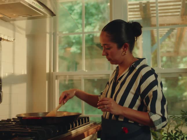 A woman picks up a spatula and stirs food in a pan boiling on the stove inside the kitchen with an apron tied around her waist