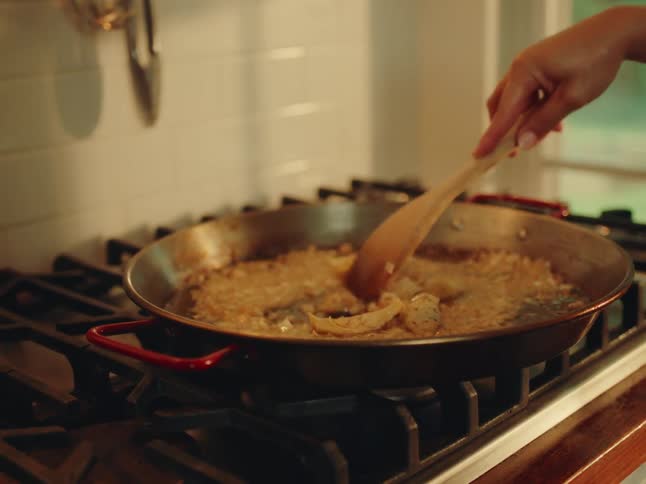 A closeup view shows a large round pan filled with simmering food possibly rice and other ingredients on a stovetop with a wooden spoon resting in it amidst a softly lit kitchen background