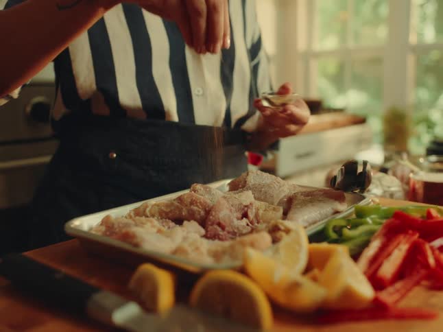 A woman in her home kitchen seasoning meat on a table that also has sliced lemons and bell peppers